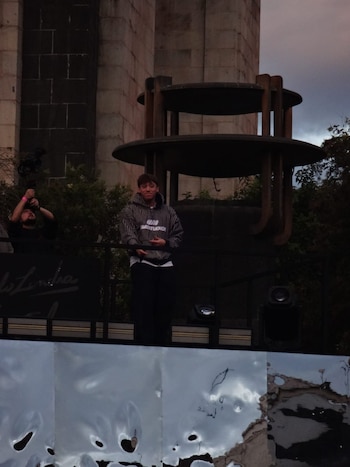 Un joven con sudadera a rayas sobre una plataforma, un camarógrafo con estabilizador a la izquierda y una estructura de piedra detrás