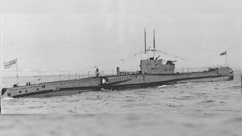 Fotografía en blanco y negro de un submarino militar gris navegando en el mar, con sus mástiles y banderas visibles