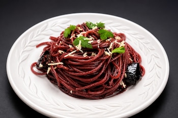 Espaguetis de color rojo oscuro con queso rallado, perejil y trozos de chile sobre un plato blanco decorado con relieve.