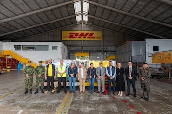 Doce personas en uniformes militares, chalecos de trabajo y trajes formales posan en fila dentro de una nave industrial con el logotipo de DHL