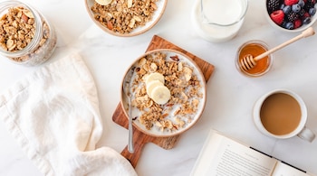 Un tazón de granola con leche y plátano, un frasco con cereales, frutos rojos, una taza de café, un frasco de miel y un libro abierto.