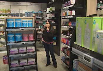 Una mujer de pie, un hombre detrás de un estante, múltiples estanterías con productos de cuidado personal, cajas de suplementos, botellas, pasillo de tienda