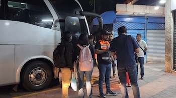 Varias personas con mochilas y un inspector con chaleco reflectante permanecen junto a un autobús blanco estacionado en la calle de noche.