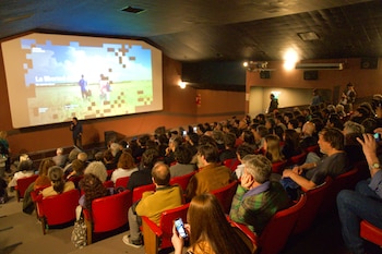 Público sentado en butacas rojas observando una pantalla gigante de proyección en una sala de cine