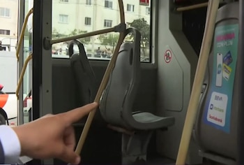 Un dedo señala la unión de un tubo vertical con un asiento gris dentro de un bus. Se observa desgaste en la estructura del vehículo, con edificios y personas por la ventana