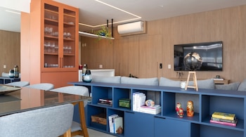 Interior moderno de sala-comedor con gabinete naranja y vidrio, estantería azul con libros y muñecas Matryoshka, televisor, sofá gris y mesa de cristal.
