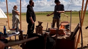 Tres personas junto a una forja de ladrillos con fuelle y yunque en un campo al aire libre bajo un toldo