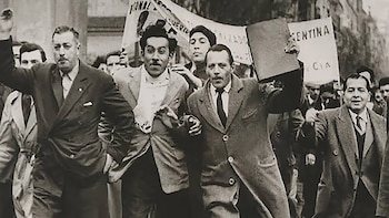 Fotografía en blanco y negro de hombres con saco y corbata marchando; uno de ellos sostiene un libro o carpeta en alto