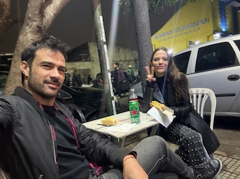 Hombre y mujer sentados en una mesa al aire libre por la noche, comen hot dogs. Hay una botella de bebida. La mujer hace la señal de la paz