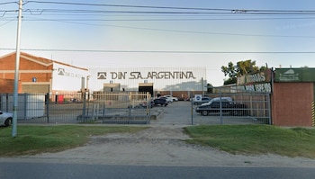 Fachada de un galpón industrial con la inscripción DIN S.A. ARGENTINA, rejas grises en el frente y vehículos estacionados