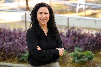 Mujer sonriente con cabello rizado oscuro, blusa negra y pantalones grises, de pie con los brazos cruzados frente a follaje borroso y una barandilla