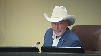 Un hombre con sombrero blanco de vaquero y saco azul se encuentra sentado frente a un micrófono en una mesa