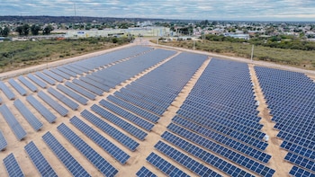 Vista aérea de un vasto campo con miles de paneles solares, caminos de tierra, edificios industriales, vegetación escasa y un pueblo al fondo