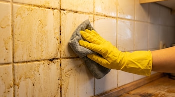 Una mano con guante de limpieza amarillo y paño gris frota una pared de azulejos de cocina sucia con grasa y manchas oscuras.