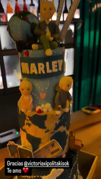 Torta de varios pisos con decoración de mapamundi en tonos azul y beige. Incluye figurines que representan a personas y el nombre 'MARLEY' en letras blancas