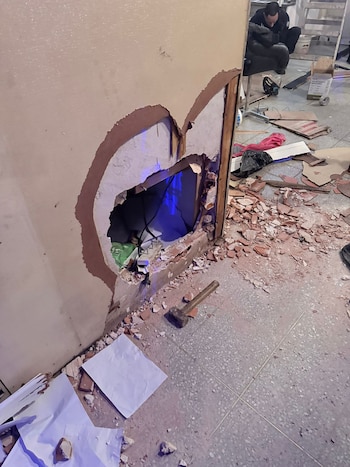 Boquete grande en una pared de color claro con escombros de ladrillos y yeso en el suelo, junto a un martillo. Un oficial se ve al fondo