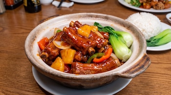 Costillas de cerdo en salsa, trozos de piña, pimientos, cebolla y bok choy en cazuela de barro. Plato con arroz blanco al fondo.