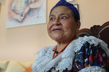 Rigoberta Menchú viste un turbante azul y un traje tradicional bordado con collar de cuentas rojas y aretes, sentada frente a una pared con un cuadro