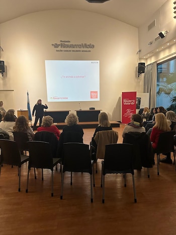 Una mujer presenta a una audiencia sentada en un salón con paredes claras y piso de madera. La pantalla proyecta un logo y la pregunta '¿Te animás a adivinar?'