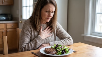 Una mujer sentada a una mesa de madera coloca su mano sobre el pecho mientras observa un plato con carne y ensalada.