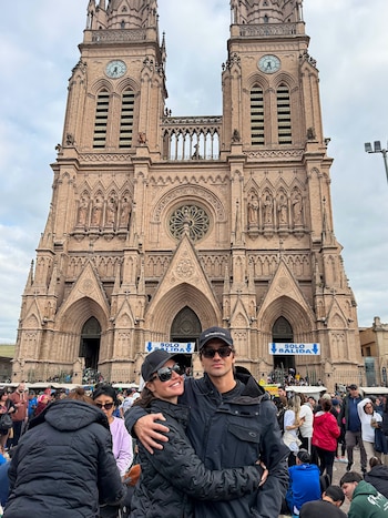 Una mujer y un hombre con gorras y gafas de sol se abrazan frente a una gran basílica neogótica rodeada de personas