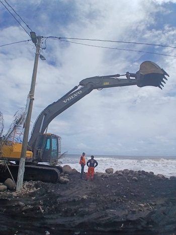Una excavadora Volvo amarilla y gris con el brazo extendido se encuentra en una playa rocosa y fangosa. Dos hombres con ropa de trabajo observan el océano agitado bajo un cielo nublado