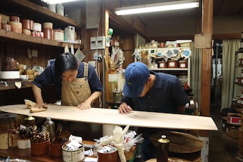 Dos hombres están inclinados sobre una tabla de madera clara dentro de un taller con estantes y recipientes