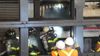 Bomberos con cascos y equipo de oxígeno. Hombres con cascos blancos y chalecos naranjas reflectantes. Humo visible. Estructura metálica