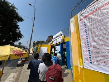 Fila de personas esperando frente a la entrada de un centro de votación de color amarillo con un cartel "BIENVENIDOS" y un panel con información electoral