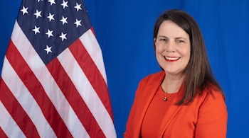 Mujer con blazer naranja y collar de perlas negras sonríe de frente, con la bandera de Estados Unidos parcialmente visible a su izquierda sobre fondo azul.