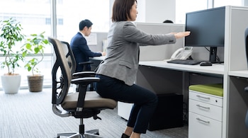 Mujer de pie frente a una silla de oficina en posición de sentadilla asistida, con los brazos extendidos. Hombre sentado en escritorio de fondo. Oficina con computadoras y plantas.