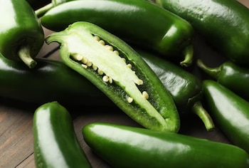 Varios pimientos jalapeños verdes sobre una superficie de madera oscura. Uno de los pimientos está cortado longitudinalmente y muestra el interior con semillas blancas