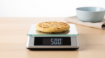 Una balanza de cocina digital con una pupusa encima; en la pantalla se lee 500. Hay una mesa de madera clara y un tazón azul con una tela detrás.