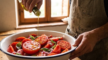 Unas manos vierten aceite de oliva sobre un recipiente blanco lleno de rodajas de tomate, cebolla y hojas de albahaca.