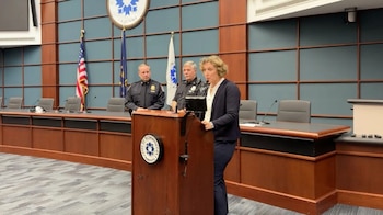 Una mujer habla desde un podio de madera con el sello de la Ciudad de Bloomington. Detrás de ella hay dos oficiales de policía y las banderas de EE.UU. e Indiana