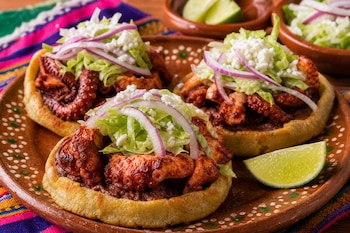 Tres sopes con tentáculos de pulpo, lechuga, cebolla y queso sobre un plato de barro con trozos de limón a un costado.