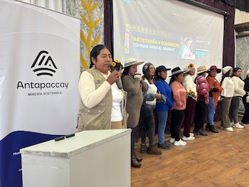 Una mujer con chaleco beige habla en un micrófono. Detrás, un banner de Antapaccay y un grupo de mujeres. Al fondo, una pantalla con texto