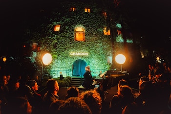 Músico tocando el piano en escenario nocturno al aire libre. Fondo: edificio con hiedra, ventanas iluminadas y logo CHANDON. Grandes farolas iluminan al público