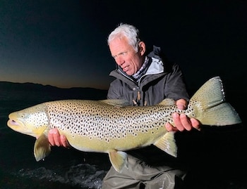 Un hombre de cabello canoso sostiene un pez grande con manchas oscuras frente a un cuerpo de agua durante la noche