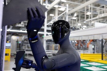 Robot humanoide de color gris oscuro con cabeza brillante negra, manipula una pieza oscura contra una superficie blanca, en una fábrica