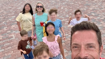Un hombre y una mujer sonríen a la cámara, rodeados de seis niños con los rostros difuminados, sobre un suelo de adoquines de tonos rojos y grises.
