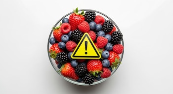 Fotografía cenital de un tazón de vidrio lleno de fresas, arándanos, moras y frambuesas, con un símbolo triangular de advertencia amarillo en el centro.