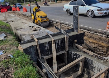 Trabajador opera compactador JCB amarillo en obra vial con zanja. Se observa hormigón fresco en encofrado de madera, barreras de tráfico naranjas y vehículos en carretera