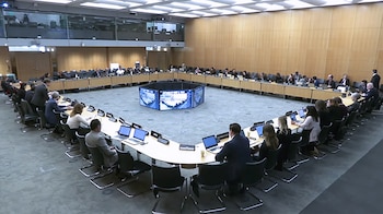 Vista aérea de una sala de conferencias con una gran mesa cuadrada y hexagonal. Numerosos delegados sentados con laptops y una pantalla central con gráficos