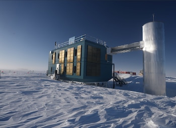 Edificio azul con paneles solares y una torre metálica conectada por un pasadizo sobre un terreno cubierto de nieve bajo un cielo despejado