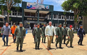 Un grupo de hombres con uniformes militares y ropa civil camina por una explanada frente a un edificio con el letrero ZODI Zulia