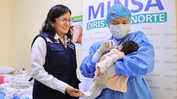Una profesional médica con mascarilla alimenta a una bebé con jeringa mientras una mujer sonriente con chaleco institucional la observa