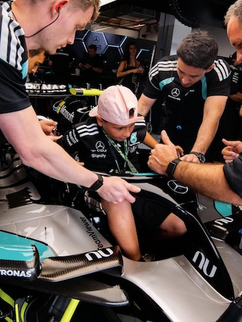 Un niño con gorra rosada y camiseta de Mercedes-AMG Petronas F1 en el asiento del piloto de un coche de F1, rodeado por personal del equipo