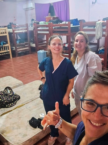 Tres mujeres sonrientes, vestidas con ropa de trabajo médica, posan para una selfie en una habitación con literas y colchones en el suelo