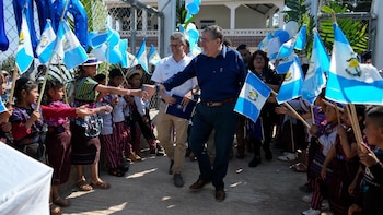 El Presidente de Guatemala, en camisa azul, saluda a niños sonrientes con banderas del país y trajes tradicionales en un evento al aire libre